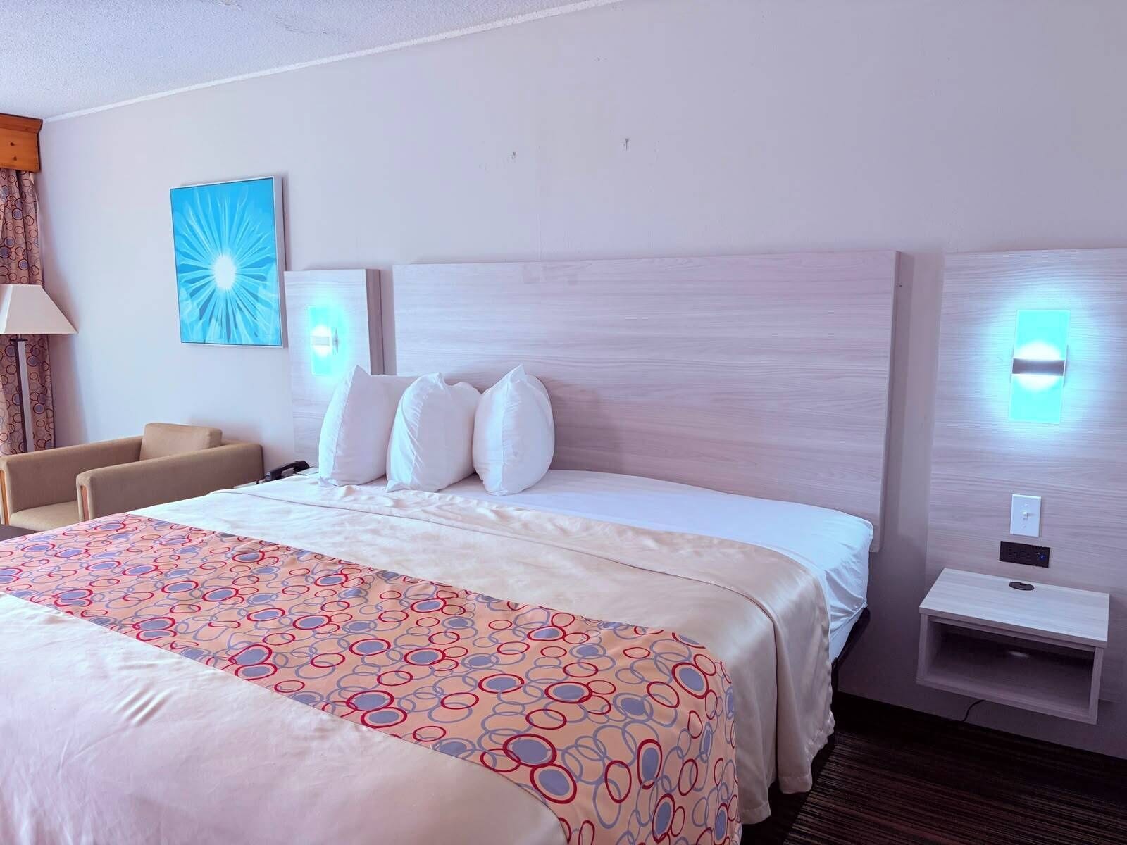 A hotel room with a large bed, a bedside table, a lamp, a chair, and a wall-mounted light fixture.