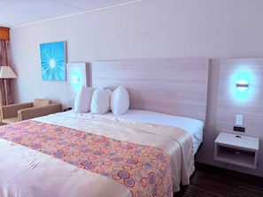 A hotel room with a large bed, a bedside table, a lamp, a chair, and a wall-mounted light fixture. - Discovery Inn Lafayette Airport I-10 (Lafayette)