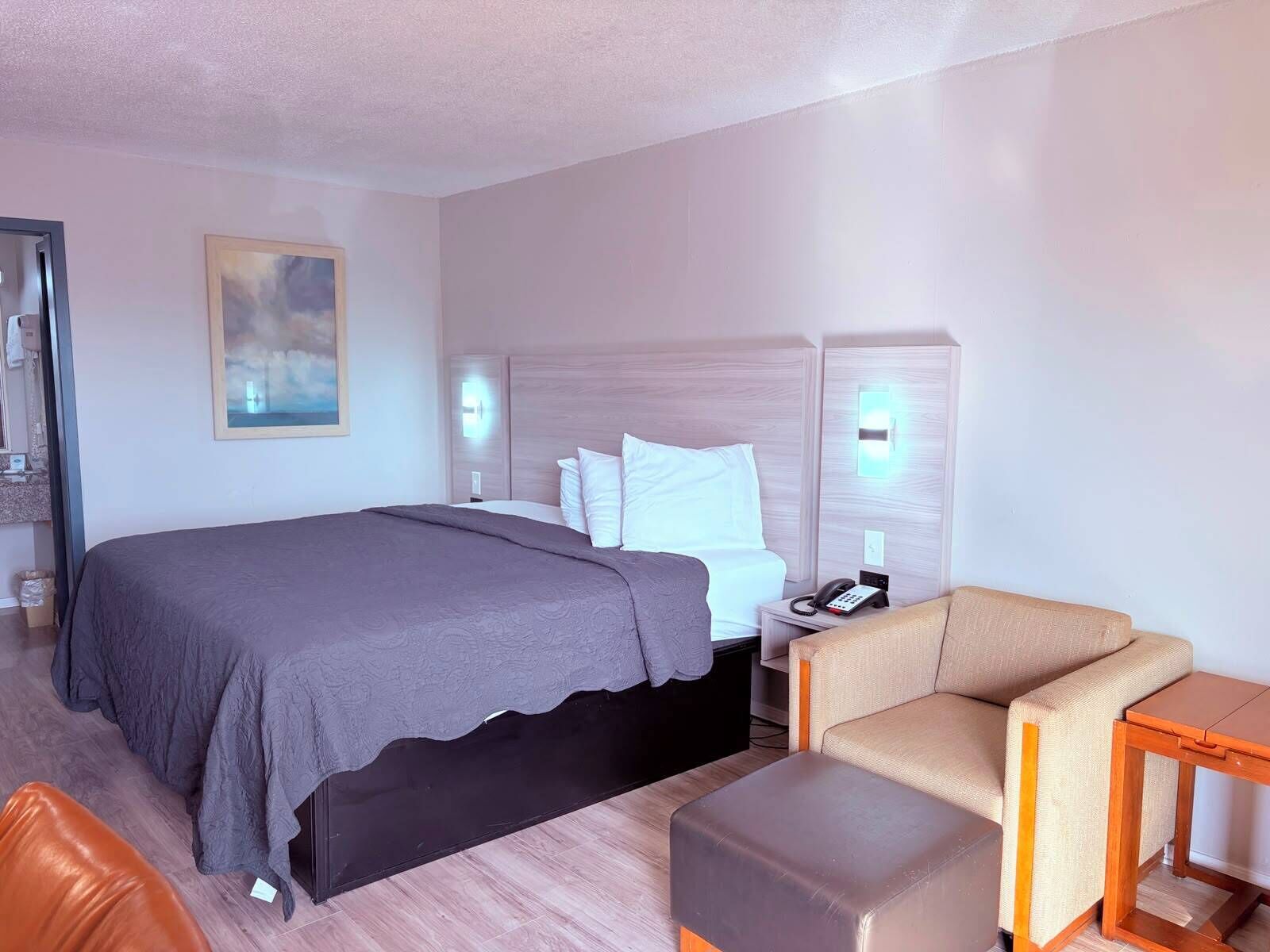 Quarto de hotel com uma cama, mesa de cabeceira com telefone, uma cadeira, uma pequena mesa e um quadro na parede.
