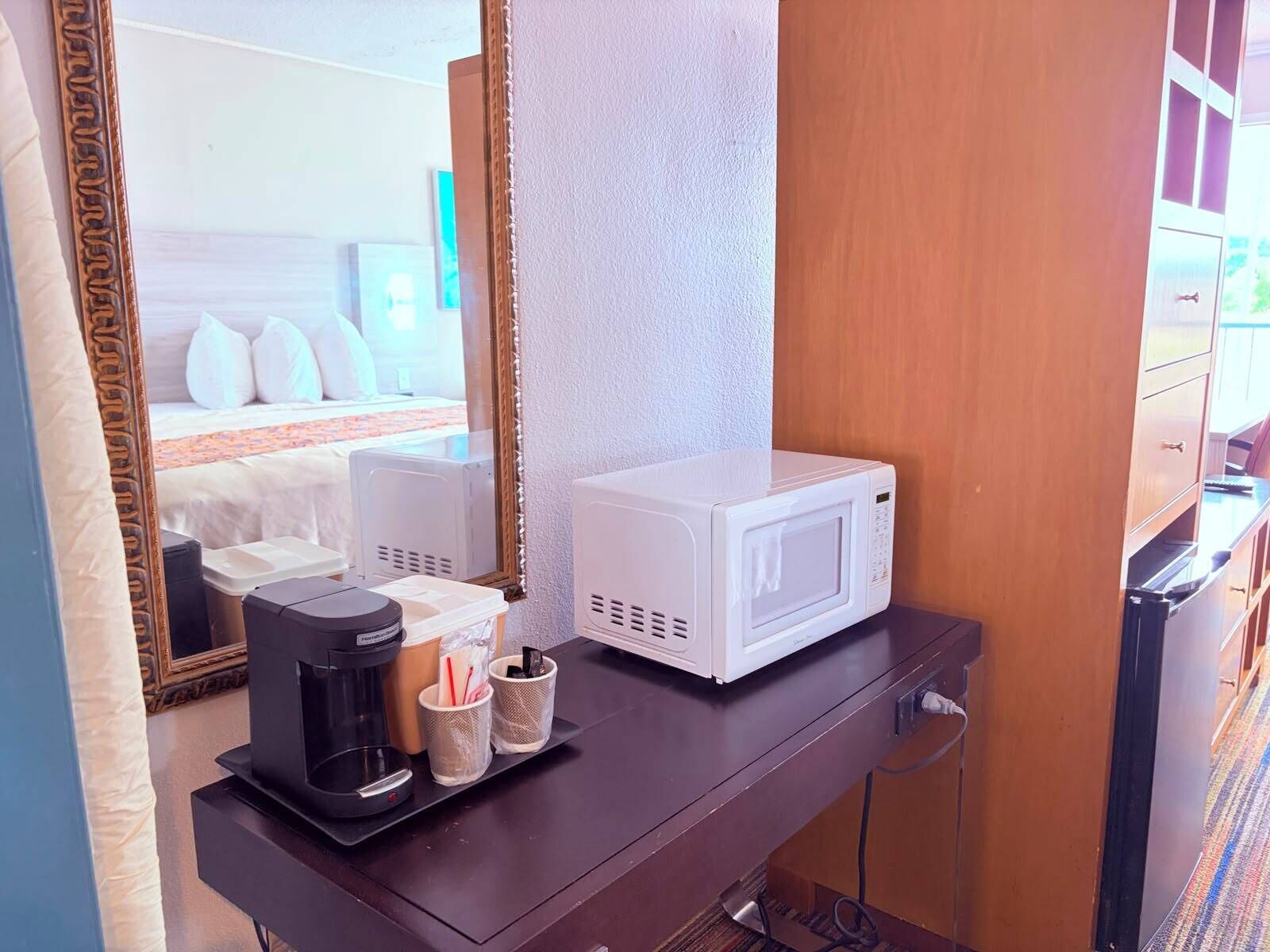 A hotel room with a bed, a white microwave on a dresser, a coffee maker, and a mirror reflecting the room.