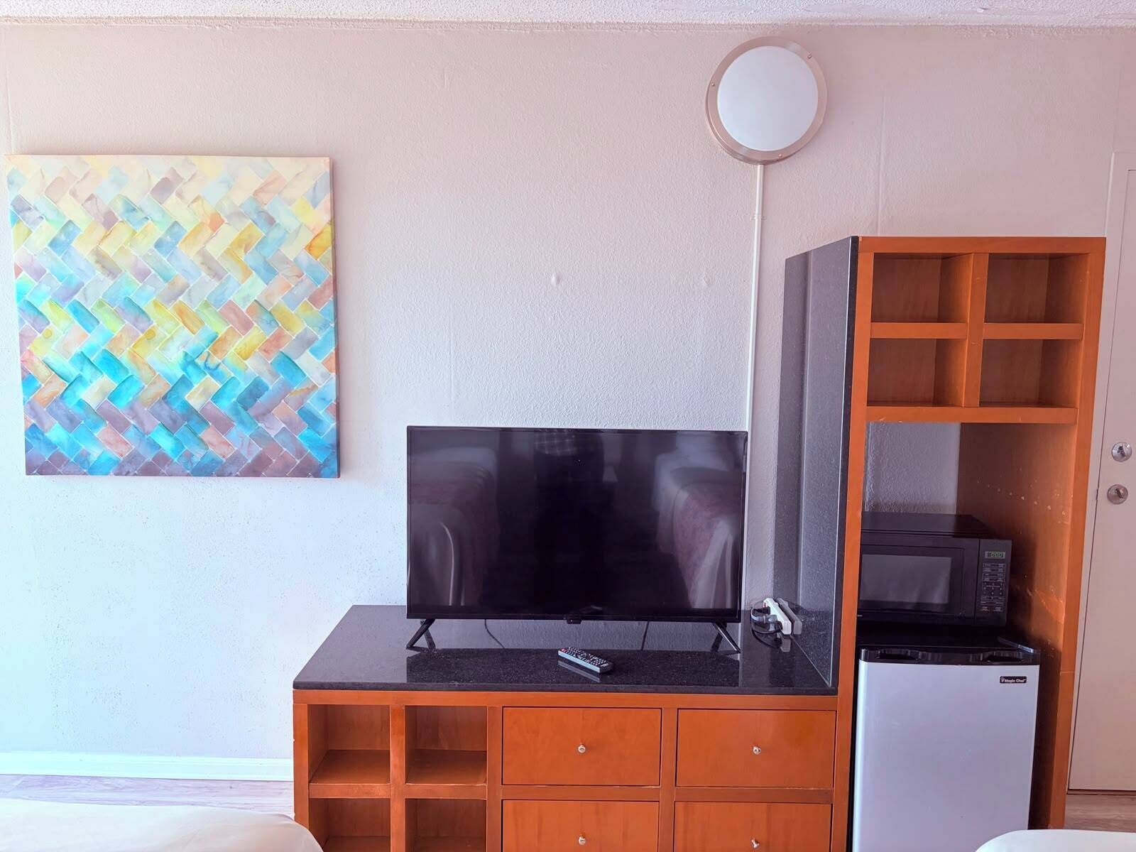 A room with a flat-screen TV on a black stand, a refrigerator, and a wooden cabinet with multiple drawers.