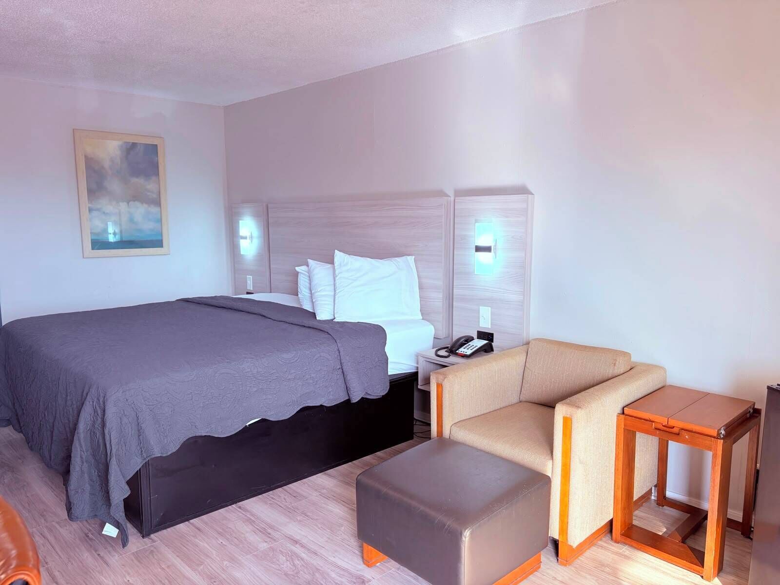 A hotel room with a bed, a chair, an ottoman, a nightstand, a phone, and a painting on the wall.