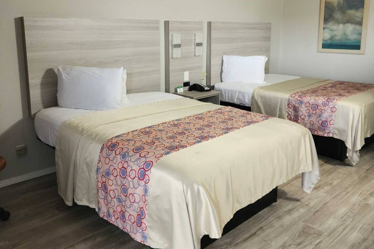 A hotel room with two beds, each with a white headboard and a patterned bedspread. There is a nightstand between the beds with a phone and a small table. The room has a wooden floor and a painting on the wall.