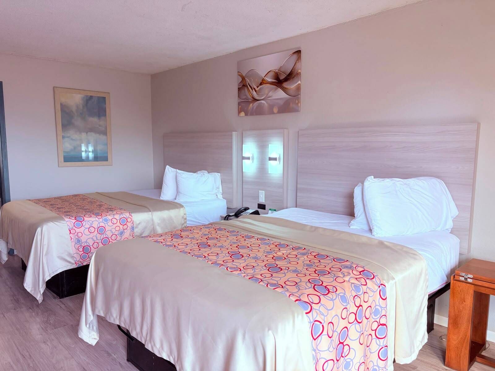 A hotel room with two beds, each with a patterned bedspread, a bedside table, a nightstand, and a wall-mounted light fixture.