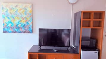 Een kamer met een televisie op een zwarte standaard, een koelkast, een houten kast met lades, en een kleurrijk abstract schilderij aan de muur.