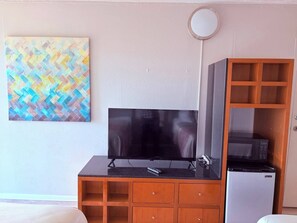 A room with a television on a black stand, a refrigerator, a wooden cabinet with drawers, and a colorful abstract painting on the wall.