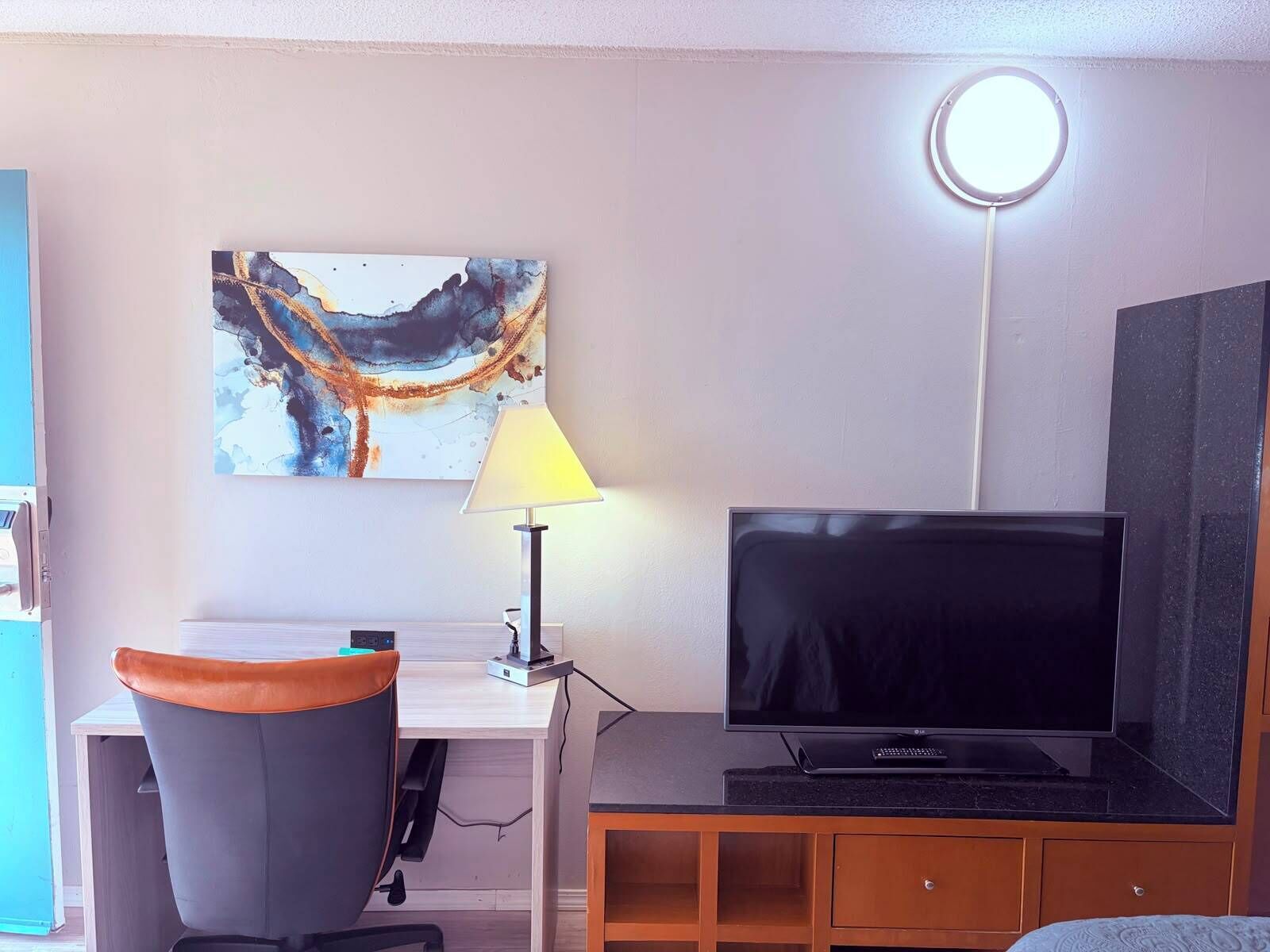 A room with a flat-screen TV, a desk with a lamp, a chair, and a wall-mounted artwork.