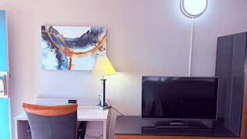 A room with a flat-screen TV, a desk with a lamp, a chair, and a wall-mounted artwork.