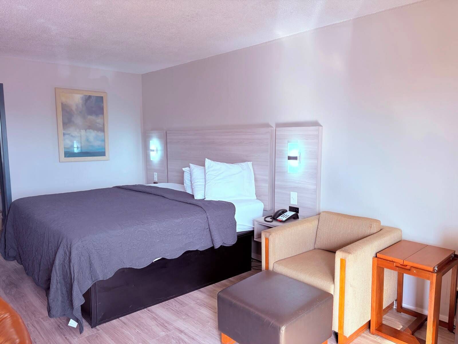 A hotel room with a bed, a chair, a small table, and a painting on the wall.