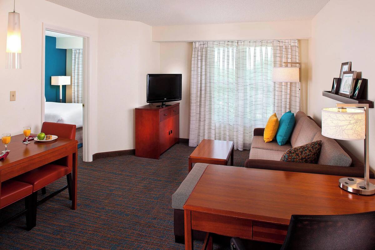 A hotel room with a wooden dining table, a sofa with colorful cushions, a TV on a wooden cabinet, and a window with patterned curtains.