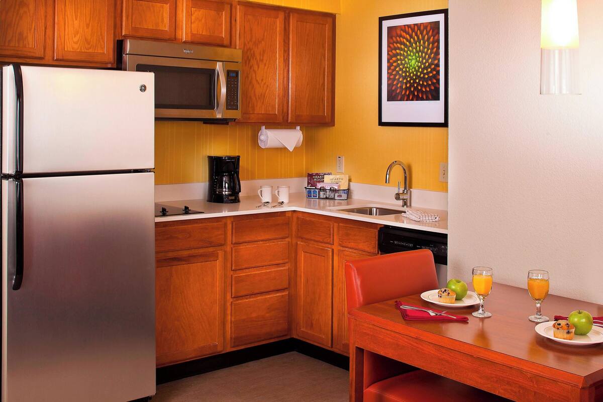 A compact kitchen with wooden cabinets, a stainless steel refrigerator, a microwave, and a coffee maker. There is a dining table with a red chair, plates with food and drinks, and a framed picture on the wall.