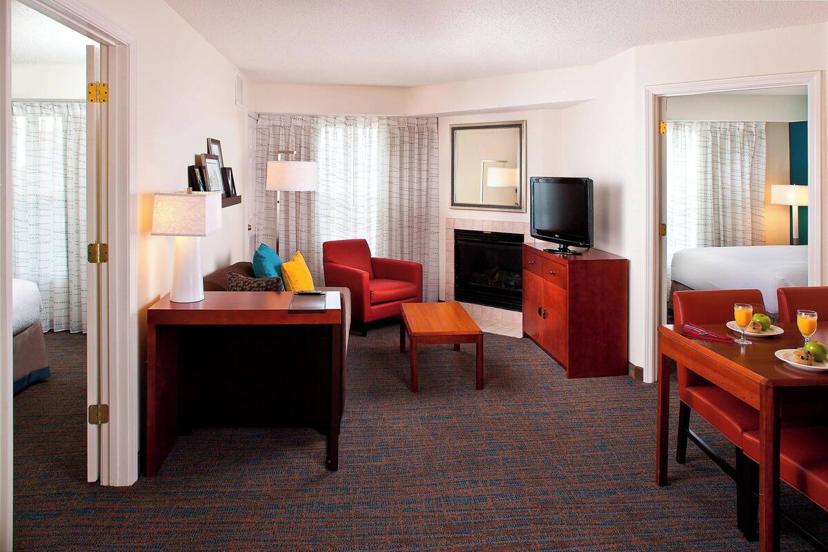 A hotel room with a dining table, a red armchair, a small wooden table, a television, and a fireplace.