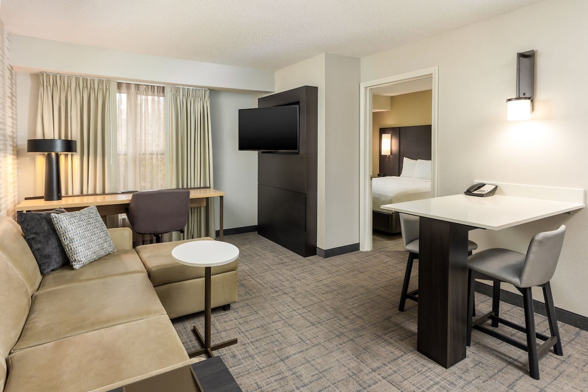 A hotel room with a sofa, a small round table, a desk with a chair, a flat-screen TV mounted on the wall, and a bed in the adjacent room.