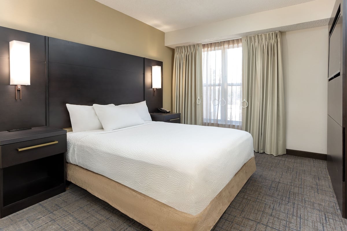 A hotel room with a large bed, dark headboard, bedside tables, a nightstand, a lamp, and a view of the outdoors through the window.