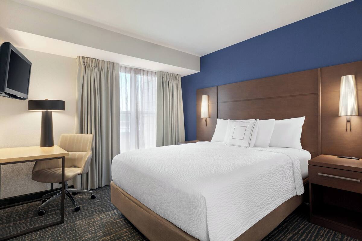 Una habitación de hotel con una cama grande, un escritorio con silla, televisión y una mesita de noche con lámpara.