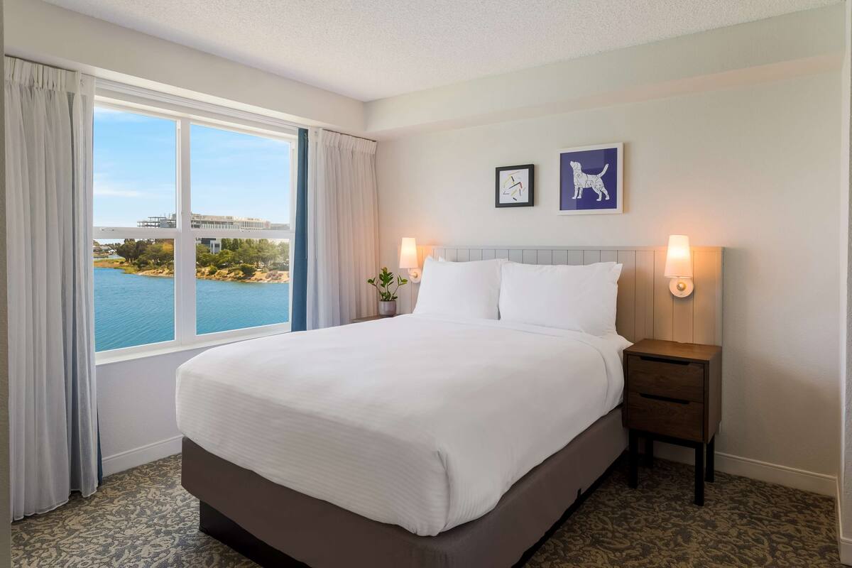 A hotel room with a large bed, two bedside lamps, a view of a body of water, and two framed pictures on the wall.