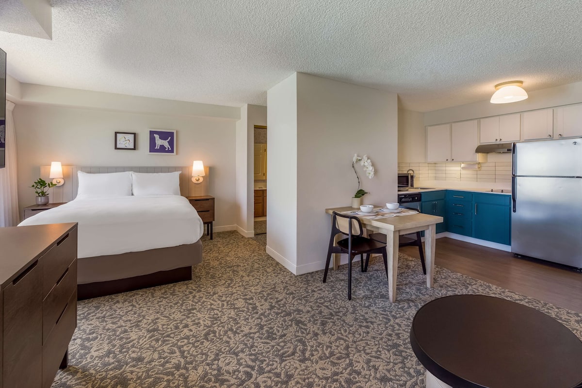 A hotel room with a bed, bedside table, lamp, and a small dining area with a table and chair.