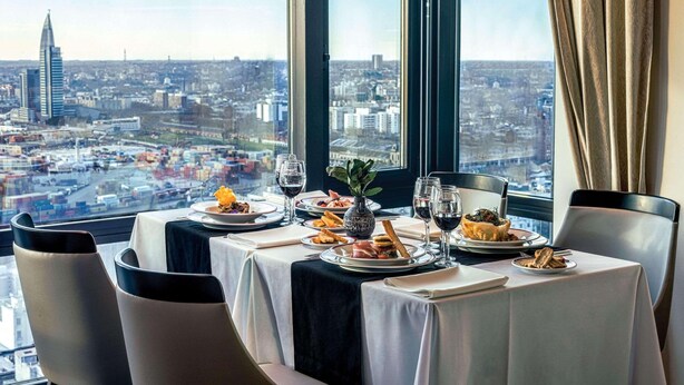 Mesa de jantar com pratos de comida e copos de vinho, com vista para a cidade através de uma grande janela.