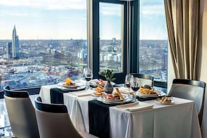 A dining table with plates of food and wine glasses, set against a cityscape view through a large window. - Radisson Montevideo Victoria Plaza Hotel (Montevideo)