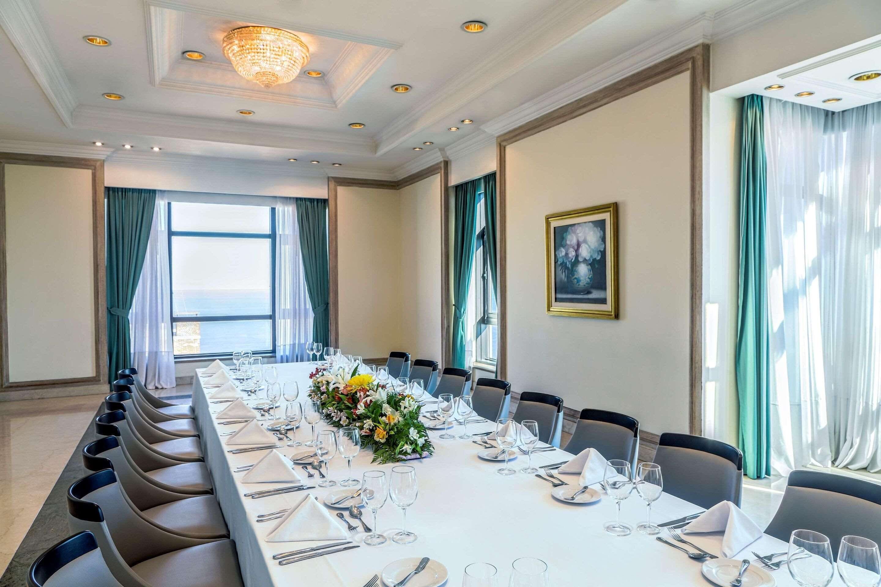A conference room with a long table set for a meeting, surrounded by chairs, with a chandelier and a painting on the wall.