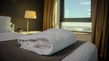 Una cama bien hecha con una toalla blanca doblada encima, una lámpara de noche y una ventana con cortinas.