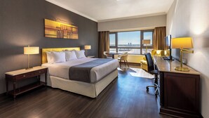 Quarto de hotel com uma cama grande, mesas de cabeceira, uma escrivaninha e uma cadeira. Há uma janela com vista para a cidade.