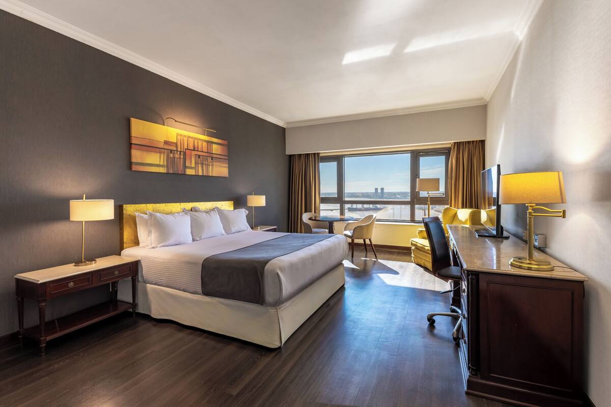 Ein Hotelzimmer mit einem großen Bett, Nachttischen, einem Schreibtisch und einem Sessel. Ein Fenster bietet Blick auf die Stadt.