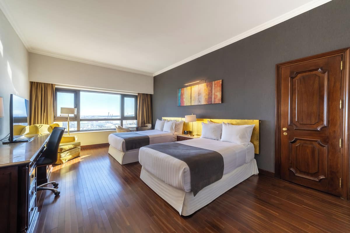 Ein Hotelzimmer mit zwei Betten, einem Schreibtisch, einem Stuhl, einer Lampe, einem großen Fenster und einer Tür.
