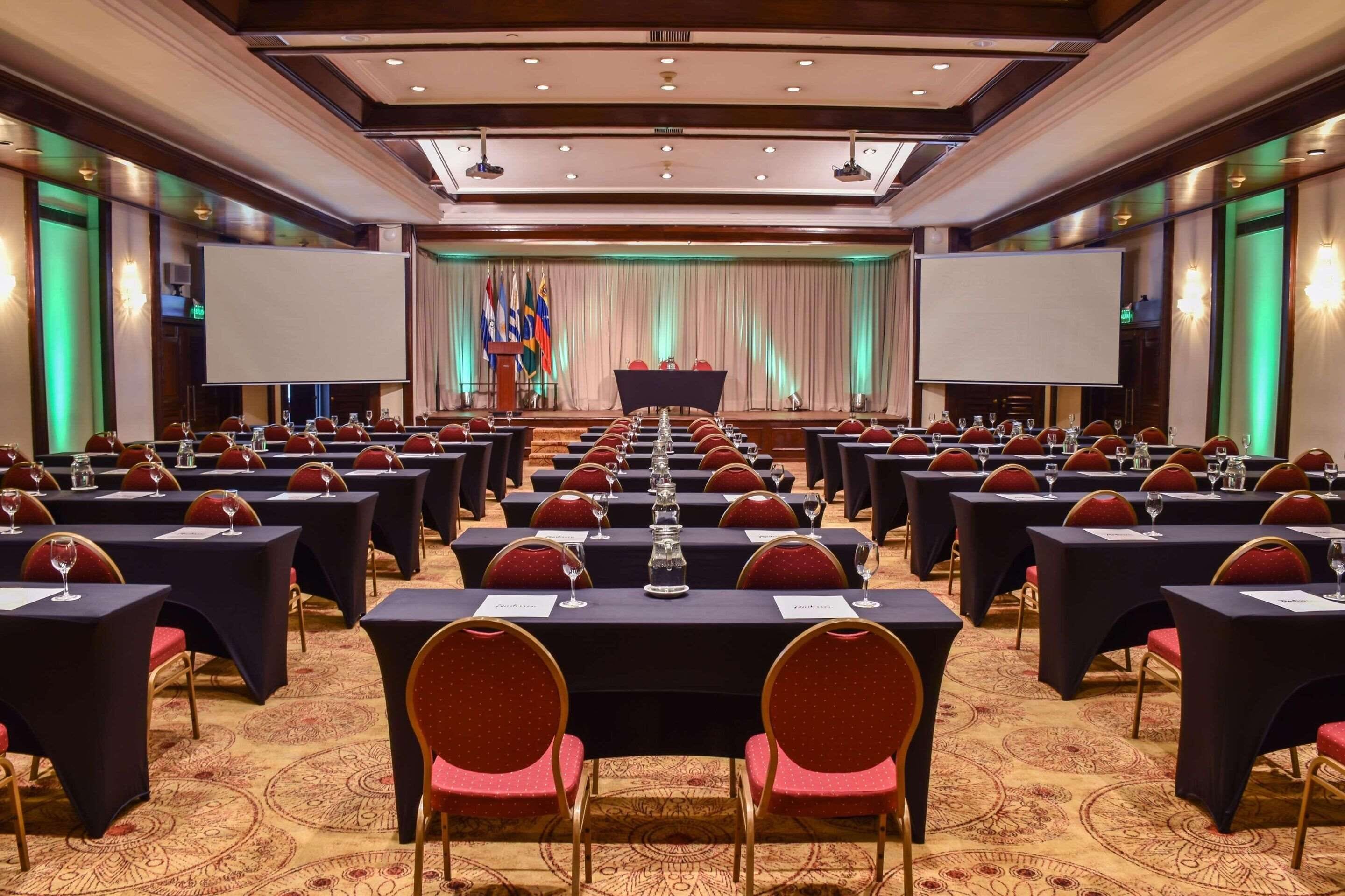A conference room with rows of tables and chairs, a stage with a podium, and flags.
