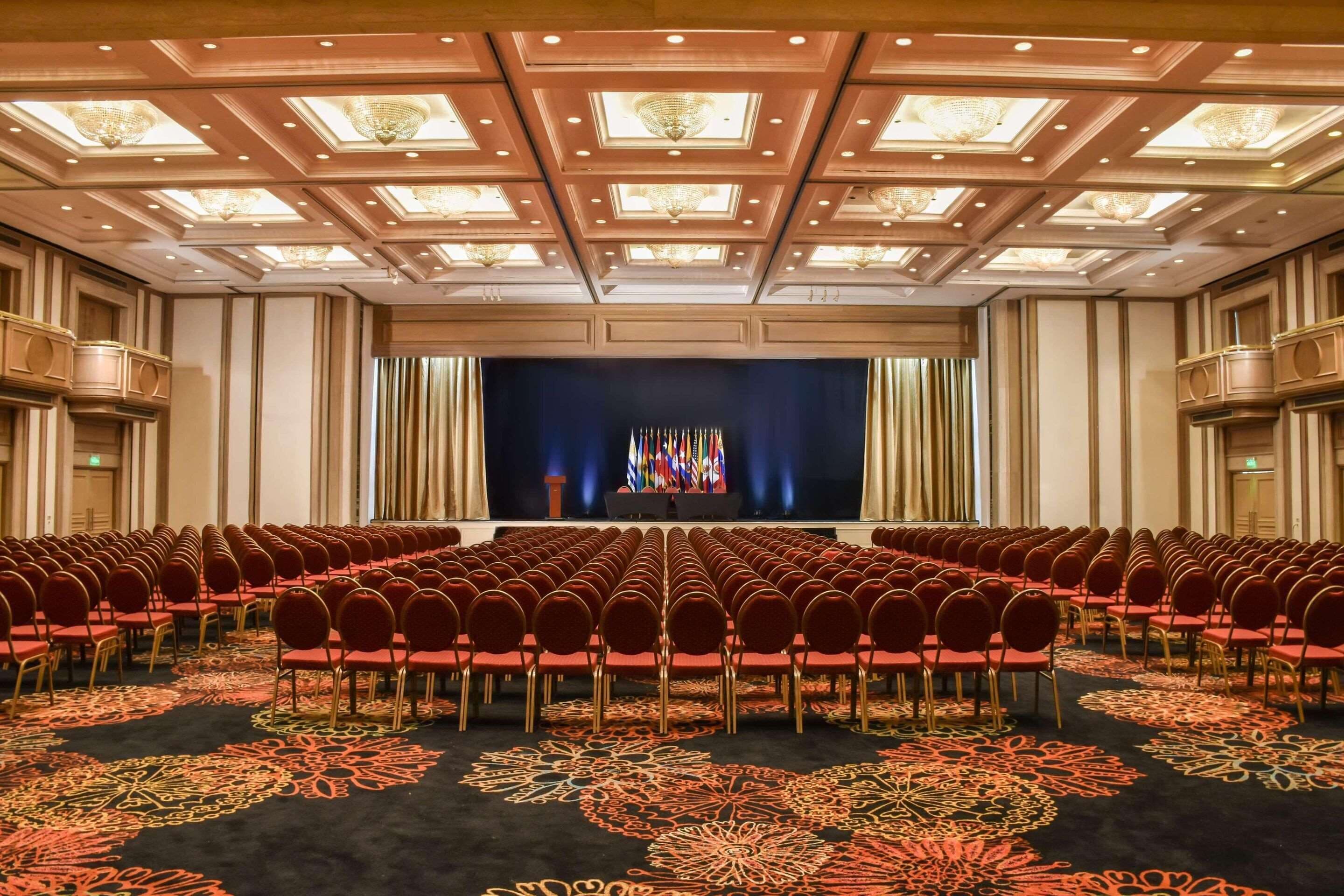 A conference room with rows of red chairs, a stage with flags, and a patterned carpet.