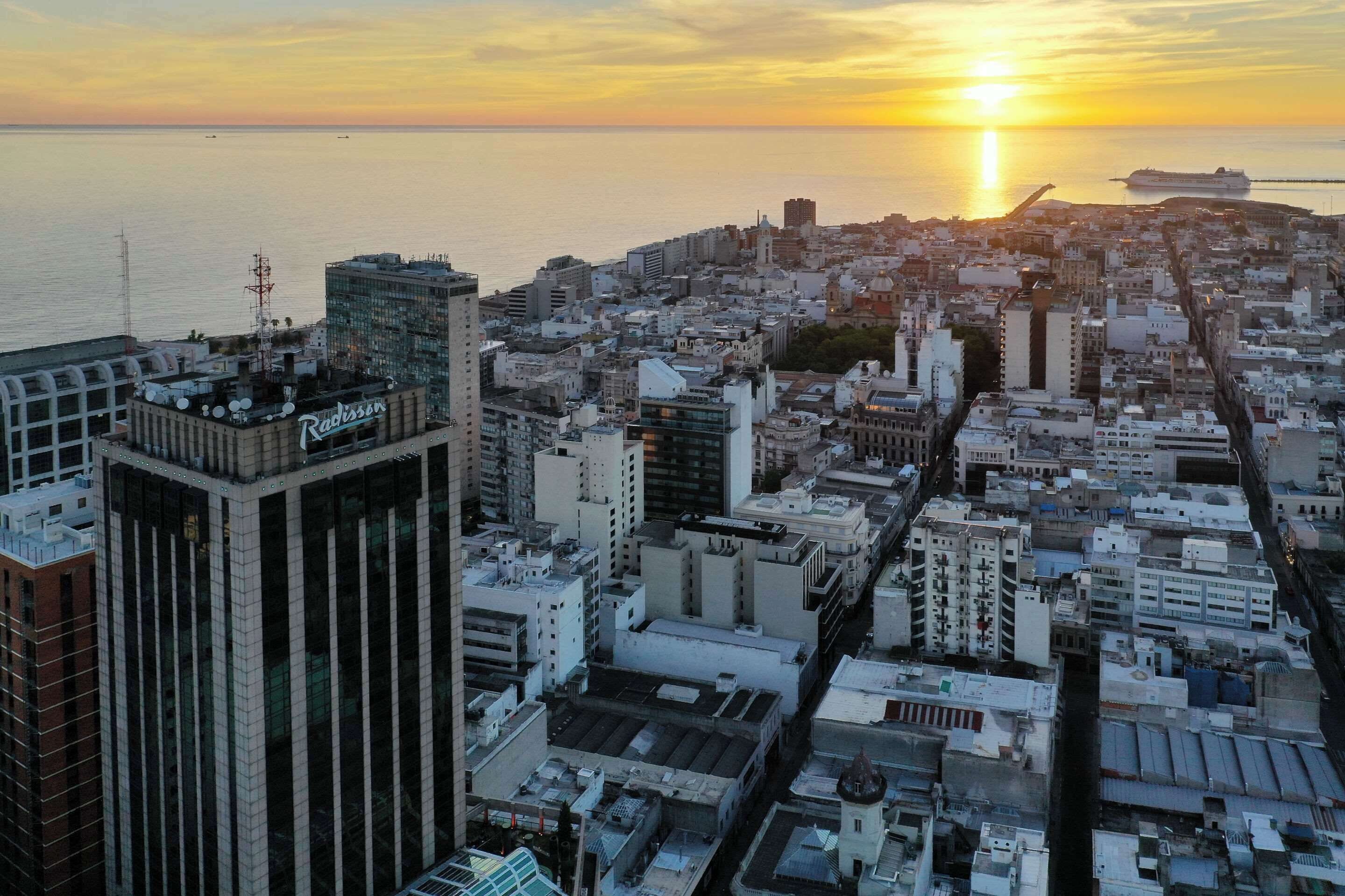 A cityscape with a prominent Radisson hotel, dense urban buildings, and a sunset over the sea.