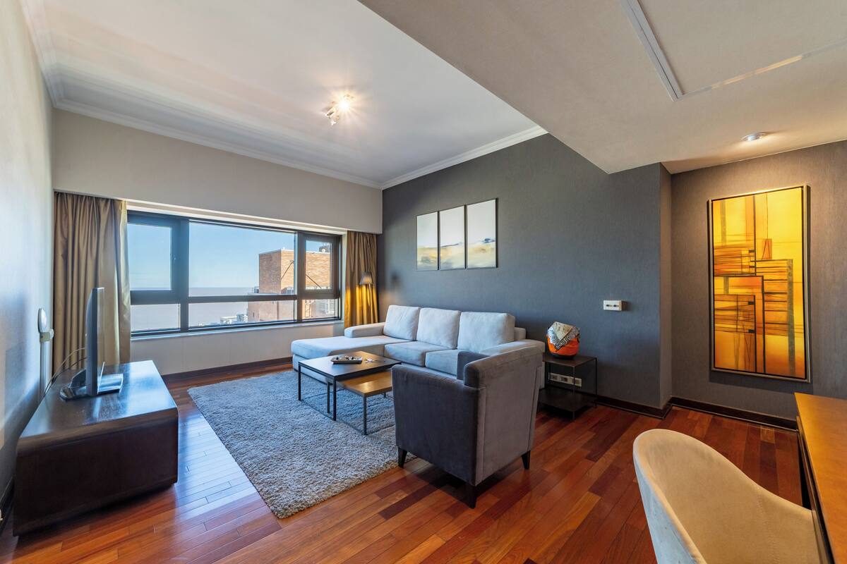 Ein modernes Wohnzimmer mit einer Couch, Sesseln, einem Couchtisch und einem Fernseher. Ein großes Fenster mit Vorhängen, ein Gemälde an der Wand und eine Lampe auf einem Beistelltisch.