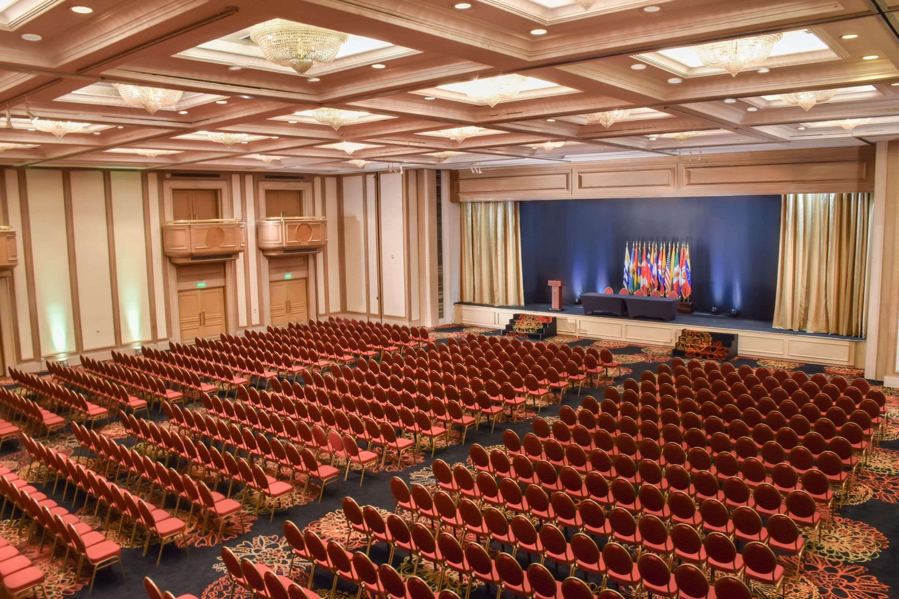 Uma sala de conferências com fileiras de cadeiras vermelhas, um palco com bandeiras e um lustre.