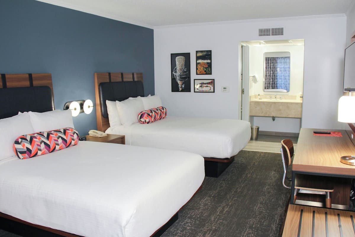 Een hotelkamer met twee bedden, een bureau, een stoel, een nachtkastje en een zichtbare badkamer door een geopende deur.