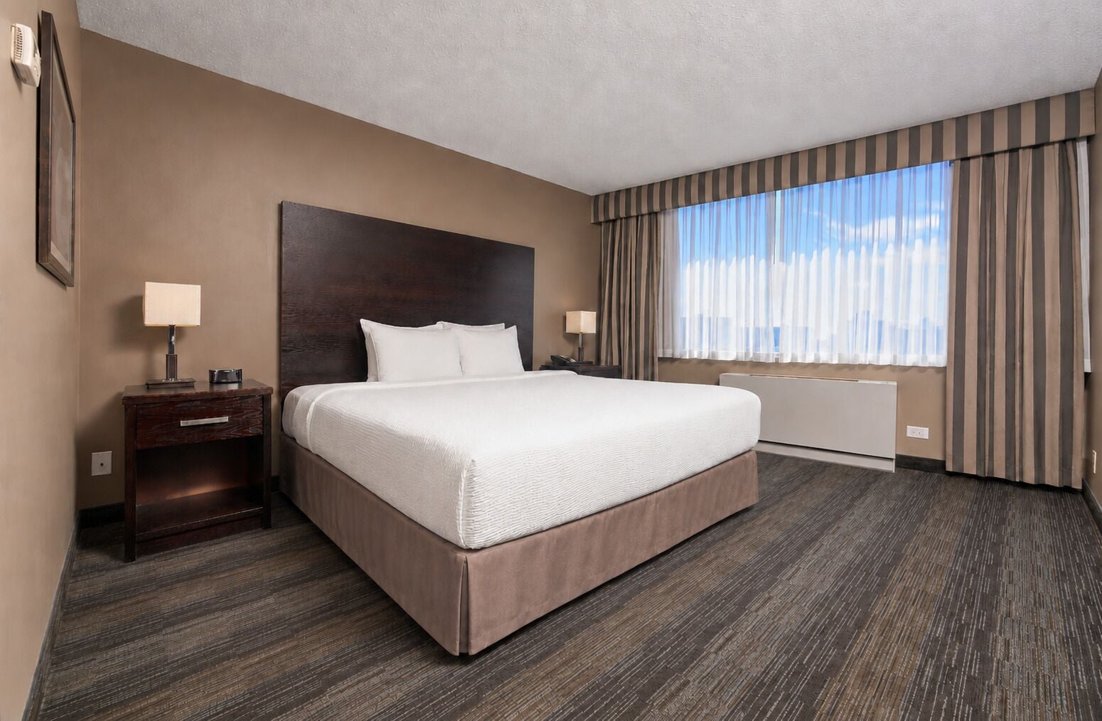 A hotel room with a large bed, bedside tables, a headboard, a window with curtains, and a radiator.