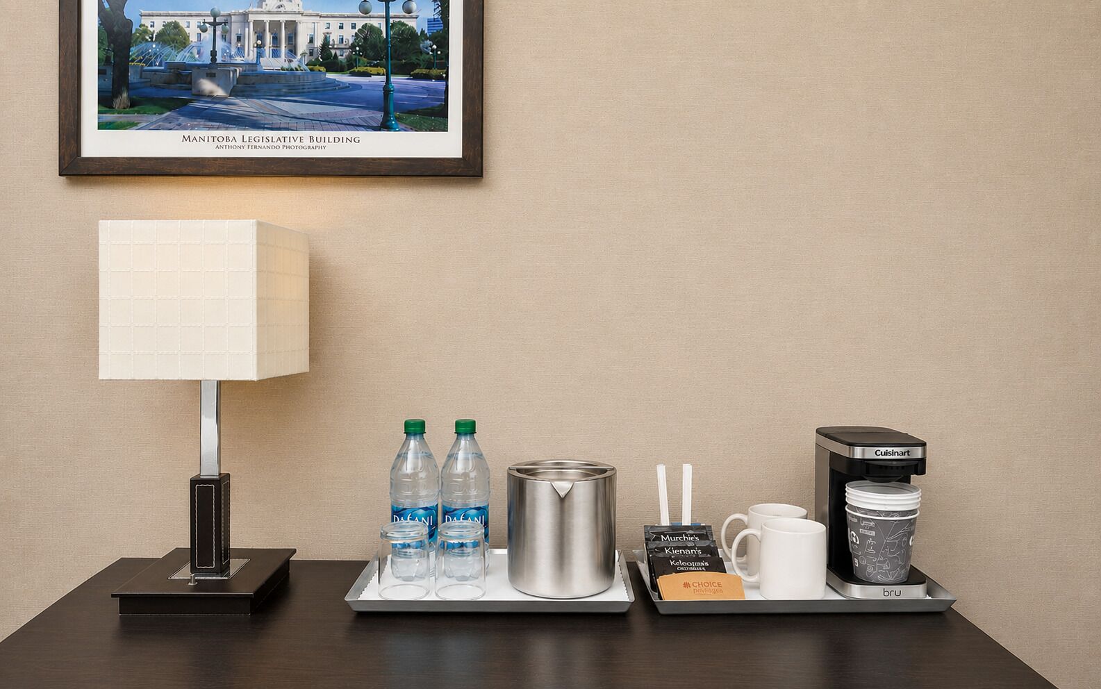 A hotel room with a framed picture of the Manitoba Legislative Building, a table with a lamp, water bottles, a thermos, a mug, a box of tea, and a coffee machine.
