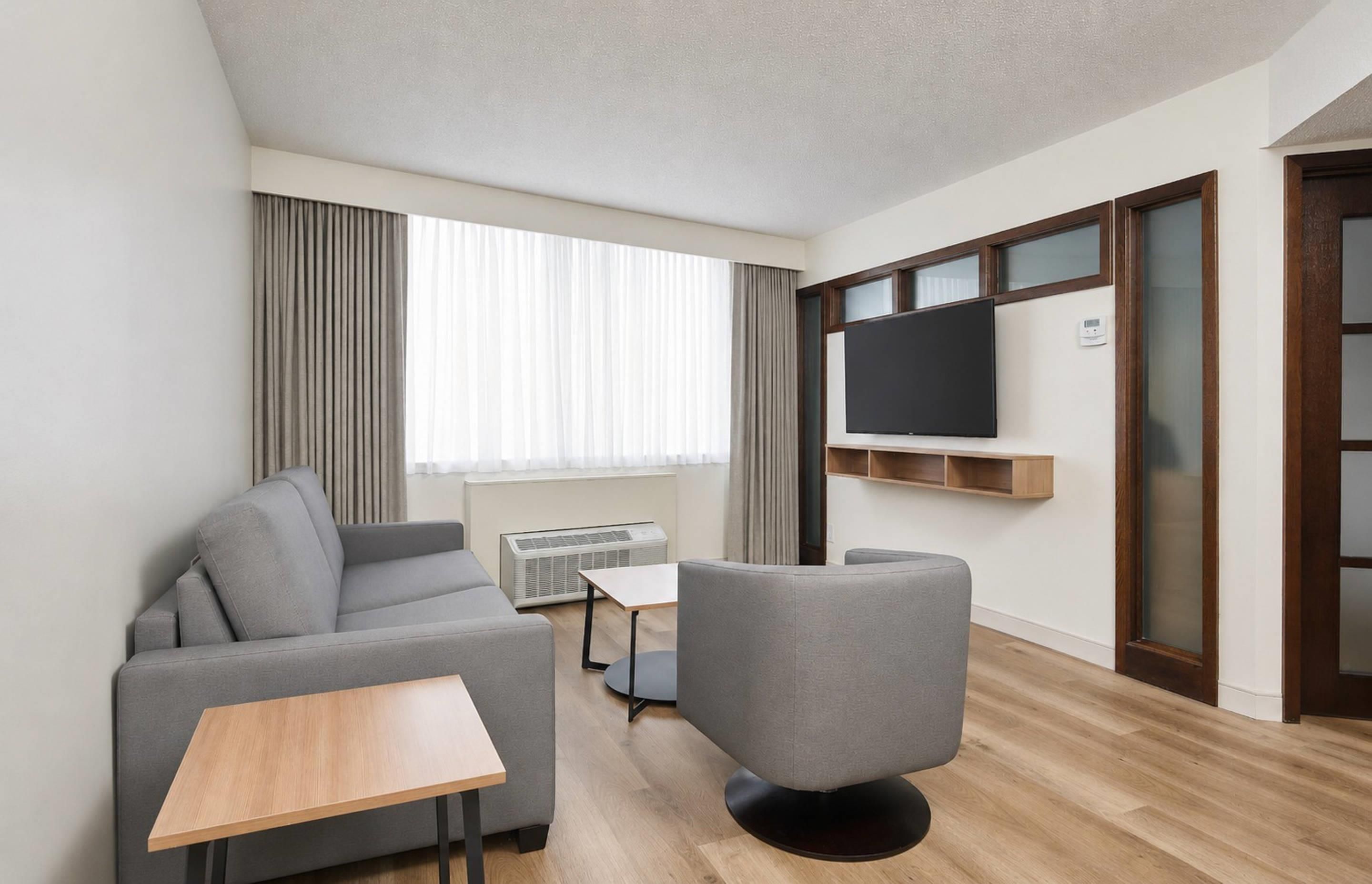 A modern living room with a grey sofa, a small table, a TV mounted on the wall, and a window with curtains.