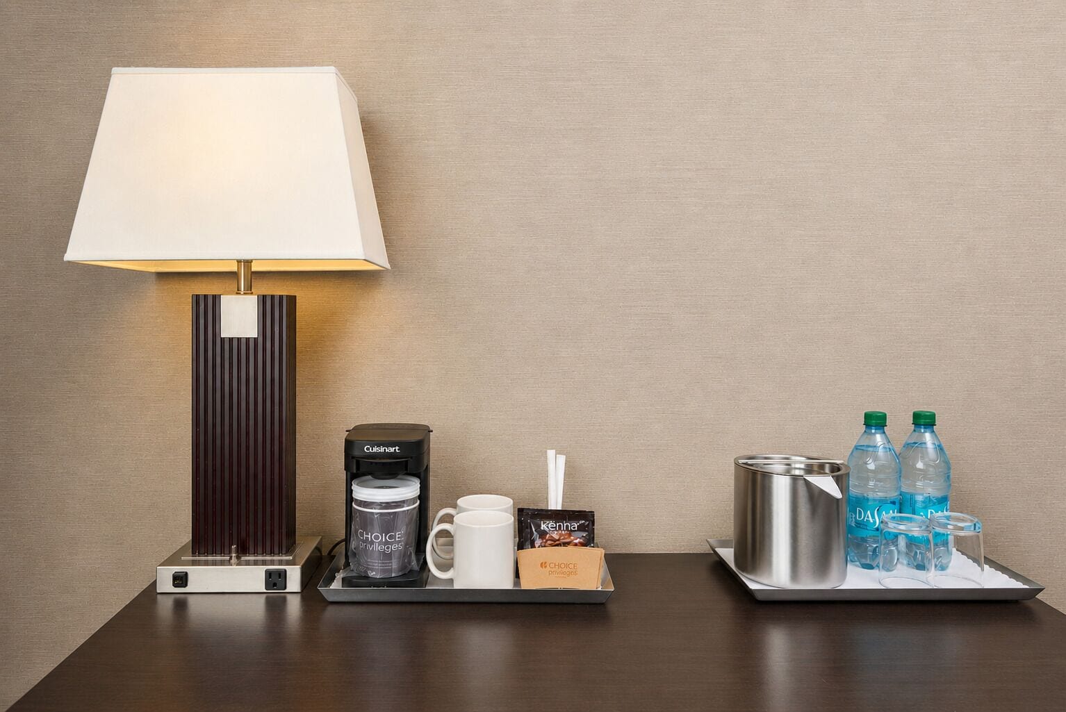 A hotel room with a bedside table featuring a lamp, a coffee maker, a tray with cups and a box of amenities, and a tray with bottled water and glasses.