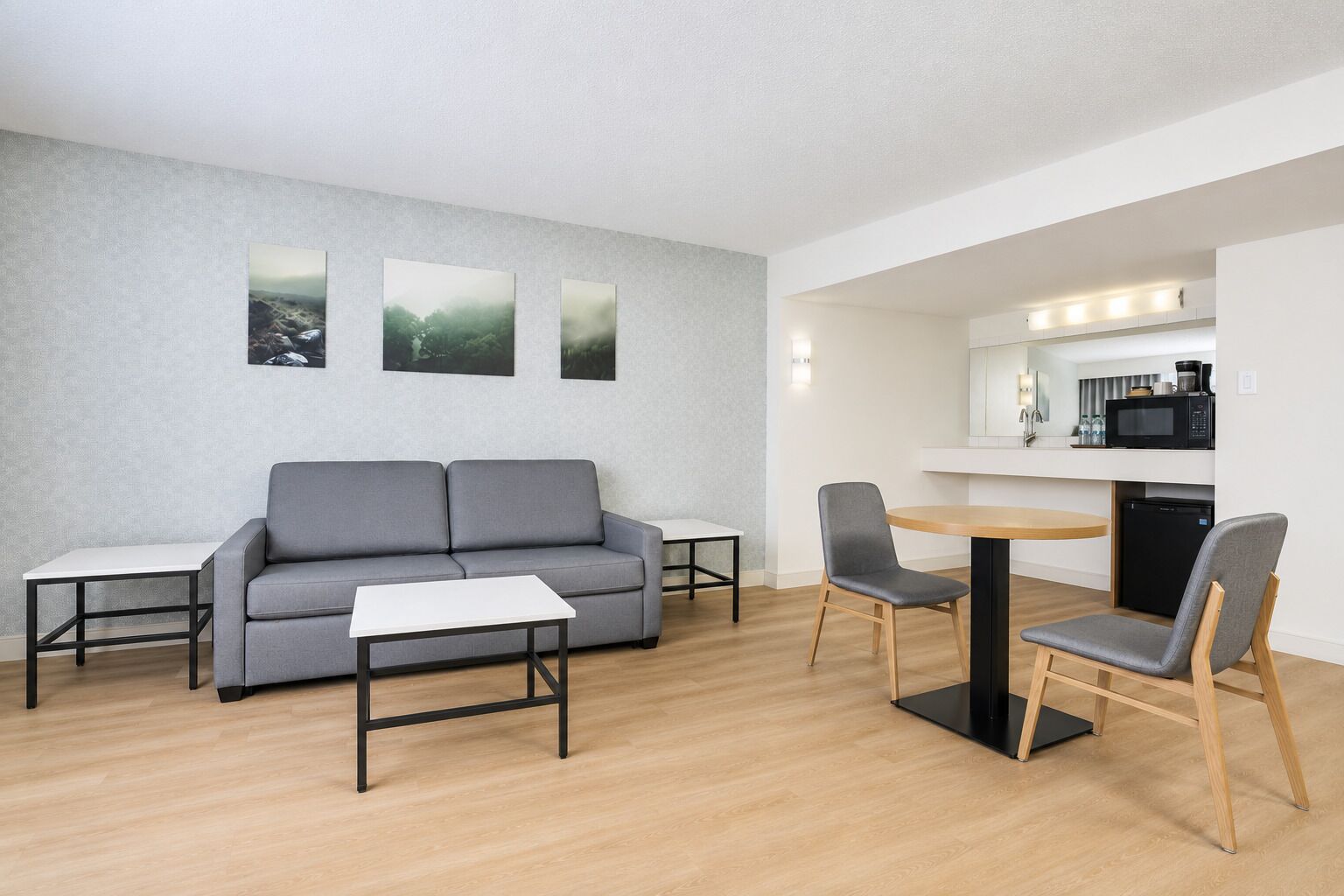A modern living room with a grey sofa, a small white table, and a round dining table with two chairs. There is a kitchen area with a microwave and a coffee maker.