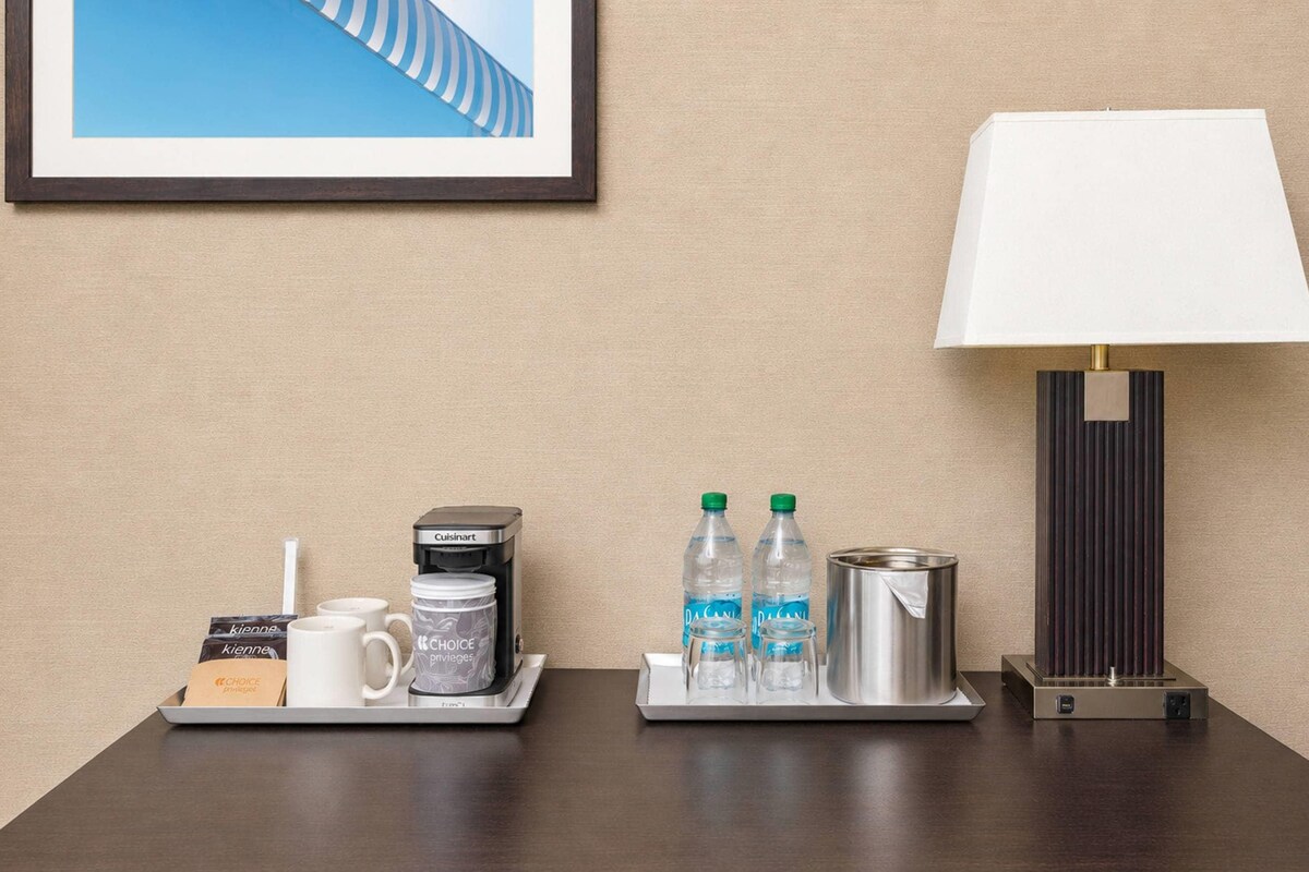A hotel room with a framed picture on the wall, a table with a tray containing a coffee maker, mugs, a water bottle, and a thermos. There is also a lamp with a white shade and a metallic base.