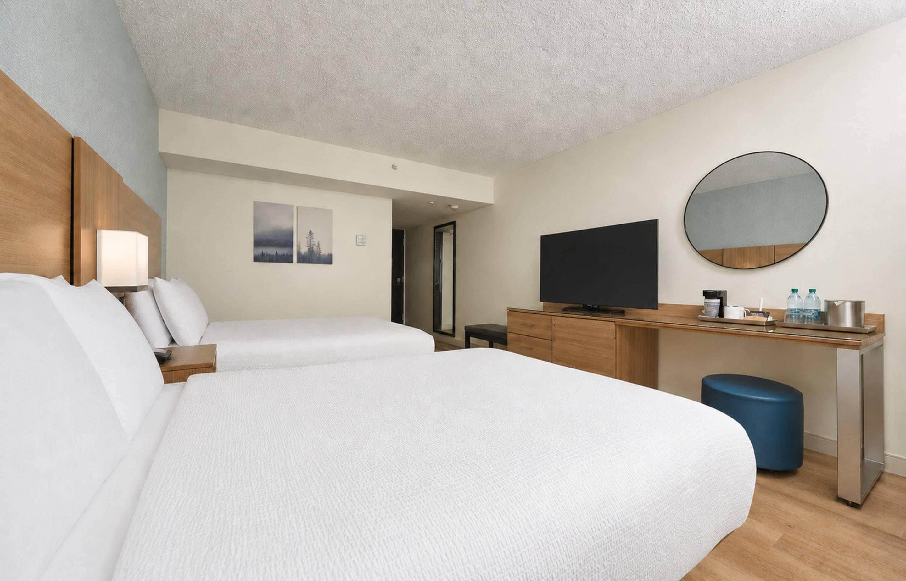 Habitación de hotel con dos camas, un escritorio, televisión de pantalla plana, un espejo y un taburete pequeño.