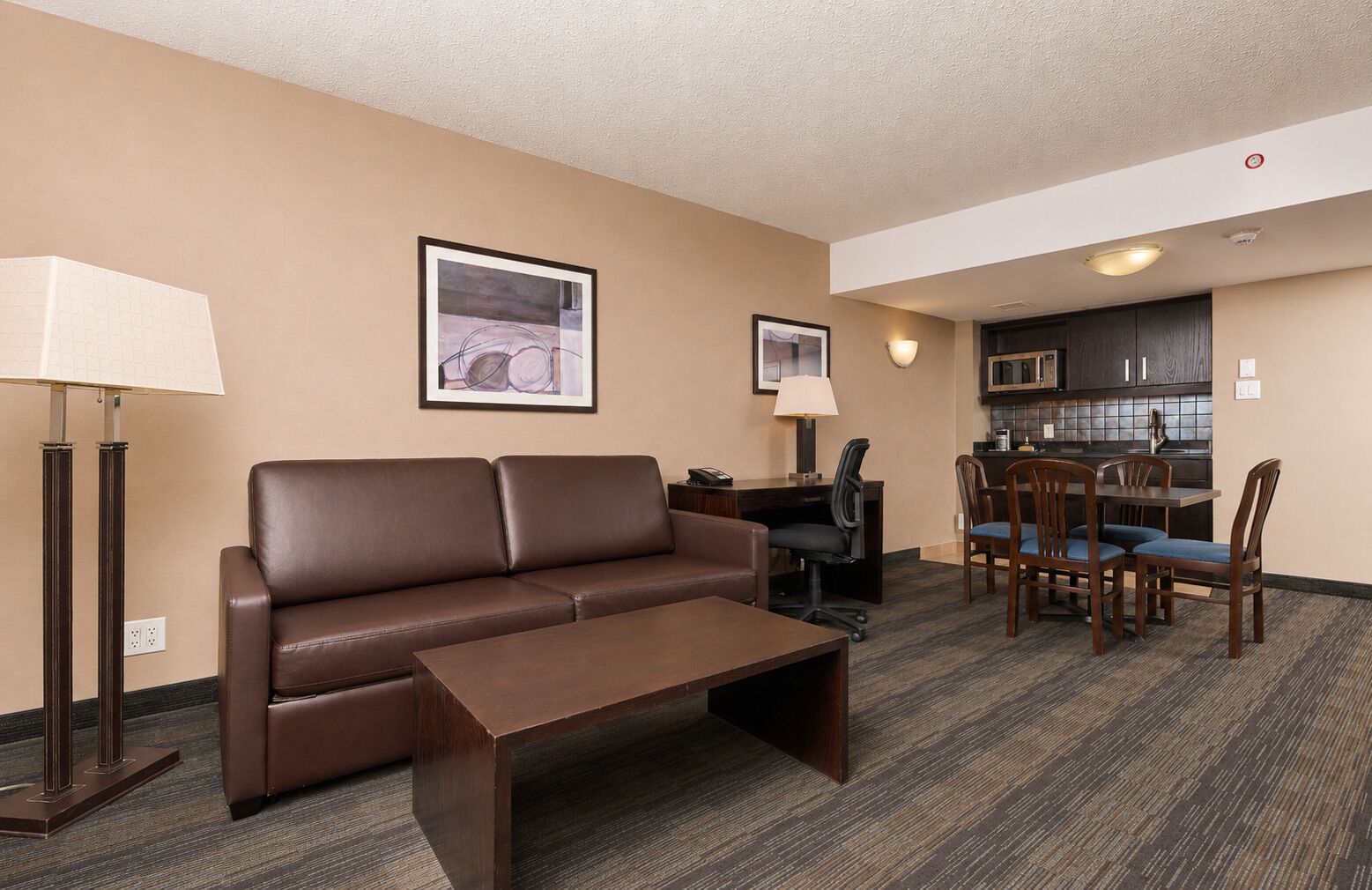 A hotel room with a sofa, a coffee table, a desk with a chair, a dining table with chairs, and a kitchenette.