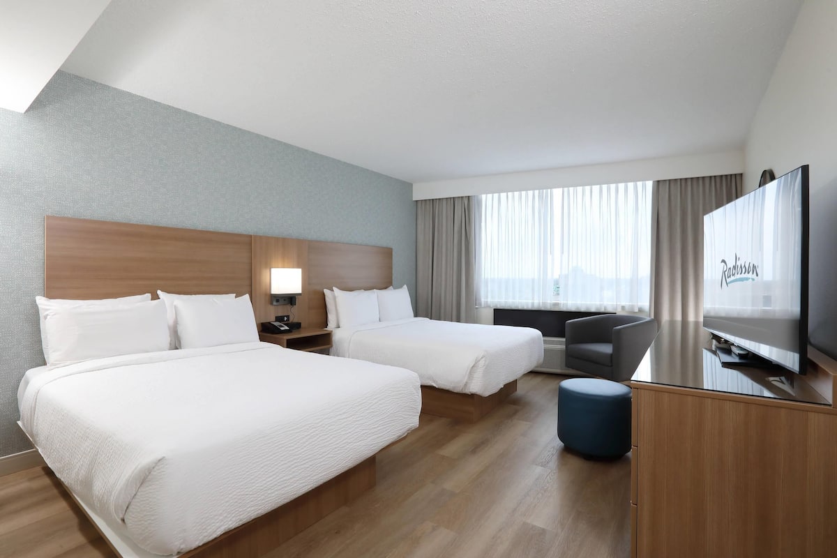 Ein Hotelzimmer mit zwei Betten, einem Schreibtisch mit Fernseher, einem Sessel und einem großen Fenster mit Vorhängen.