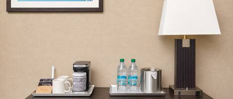 A hotel room with a framed picture on the wall, a bedside table with a lamp, a tray with a coffee maker, water bottles, and a container of coffee creamer.