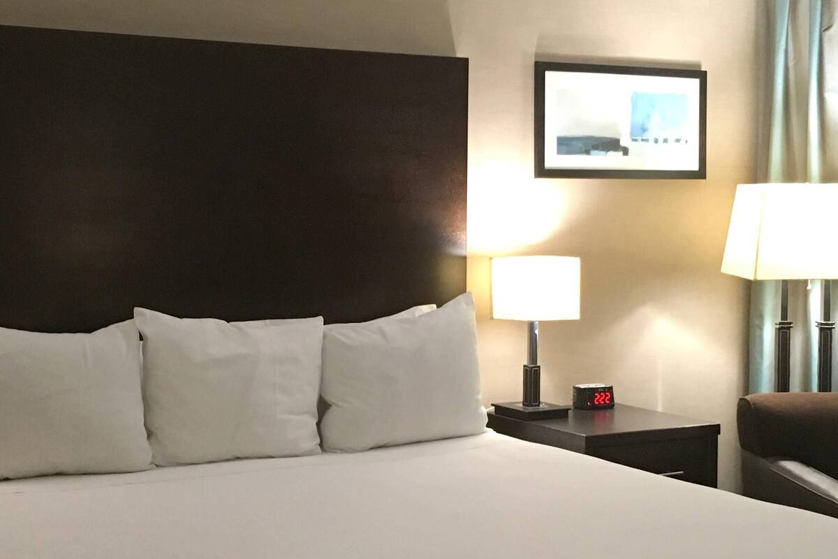 Ein Hotelzimmer mit einem großen Bett, zwei Nachttischen, einer Nachttischlampe, einer digitalen Uhr und einem gerahmten Bild an der Wand.