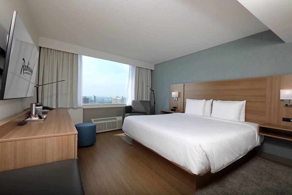 Ein Hotelzimmer mit einem großen Bett, einem Schreibtisch, einem Stuhl, einer Lampe und Blick auf die Stadt.