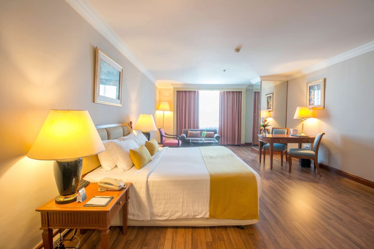 Habitación de hotel con una cama grande, lámparas de noche, un escritorio con silla y vistas al exterior a través de una ventana.