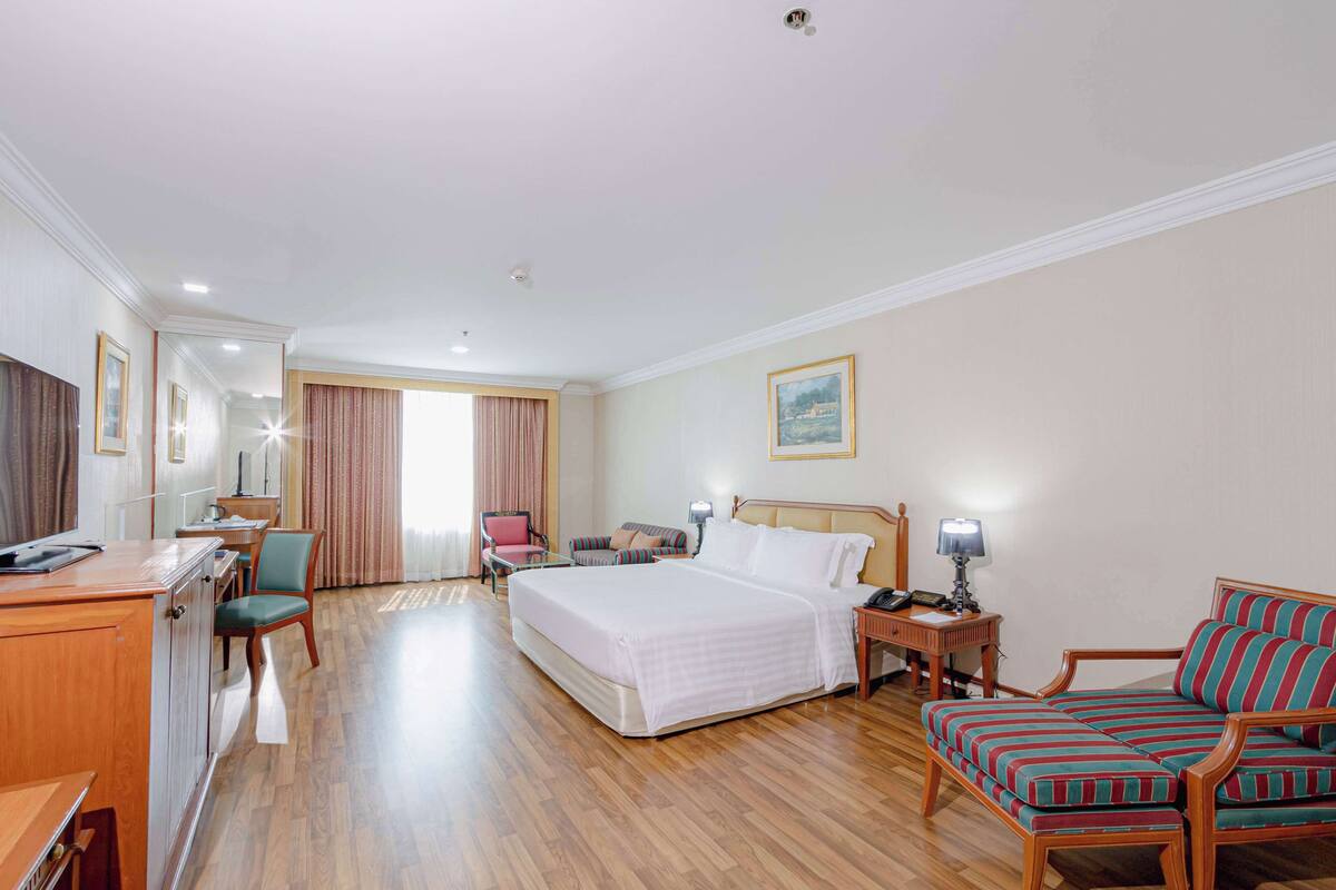 A hotel room with a bed, a desk with a chair, a sofa, and a small table with a lamp.