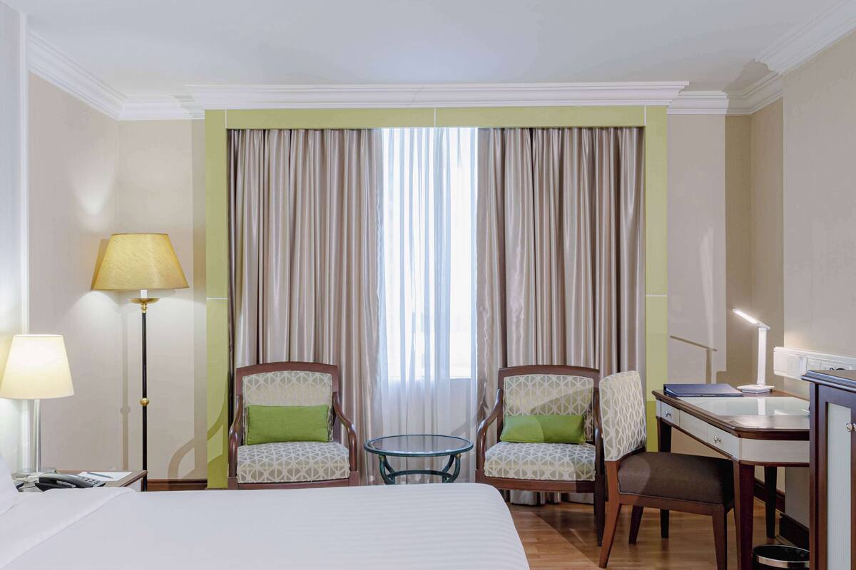 A hotel room with a bed, two armchairs with green cushions, a small round table, a desk with a lamp, and a window with curtains.