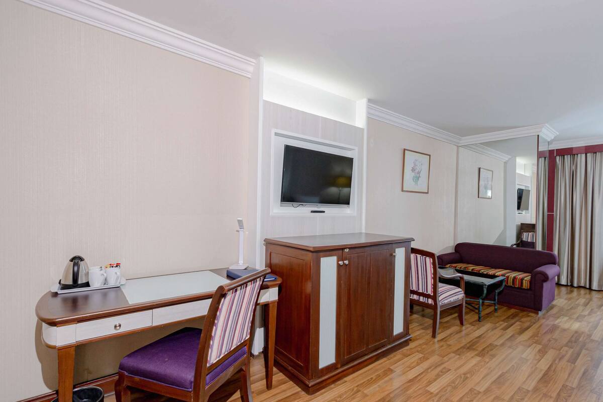 A hotel room with a wooden desk, a purple chair, a sofa, and a television mounted on the wall.